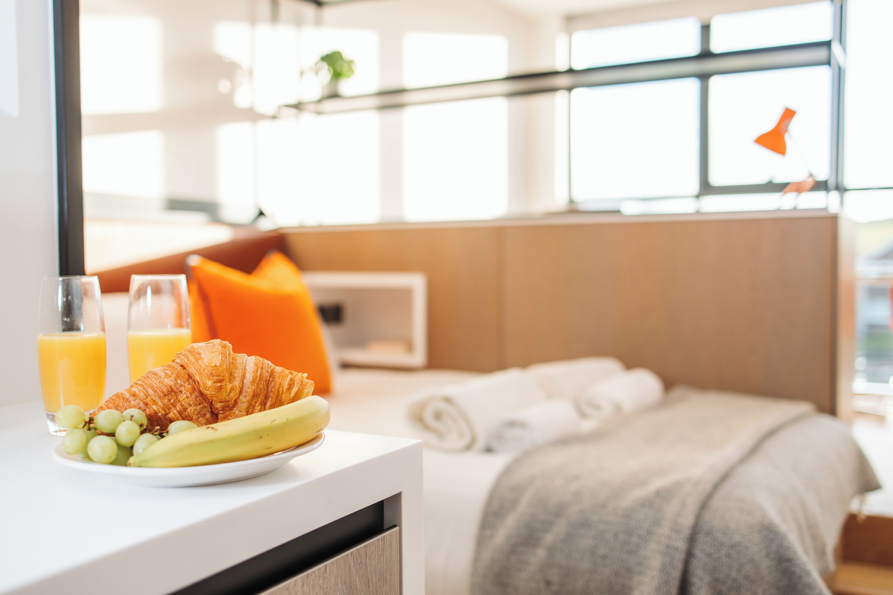 A hotel room with a bed, a bedside table with a croissant, grapes, a banana, and two glasses of orange juice.