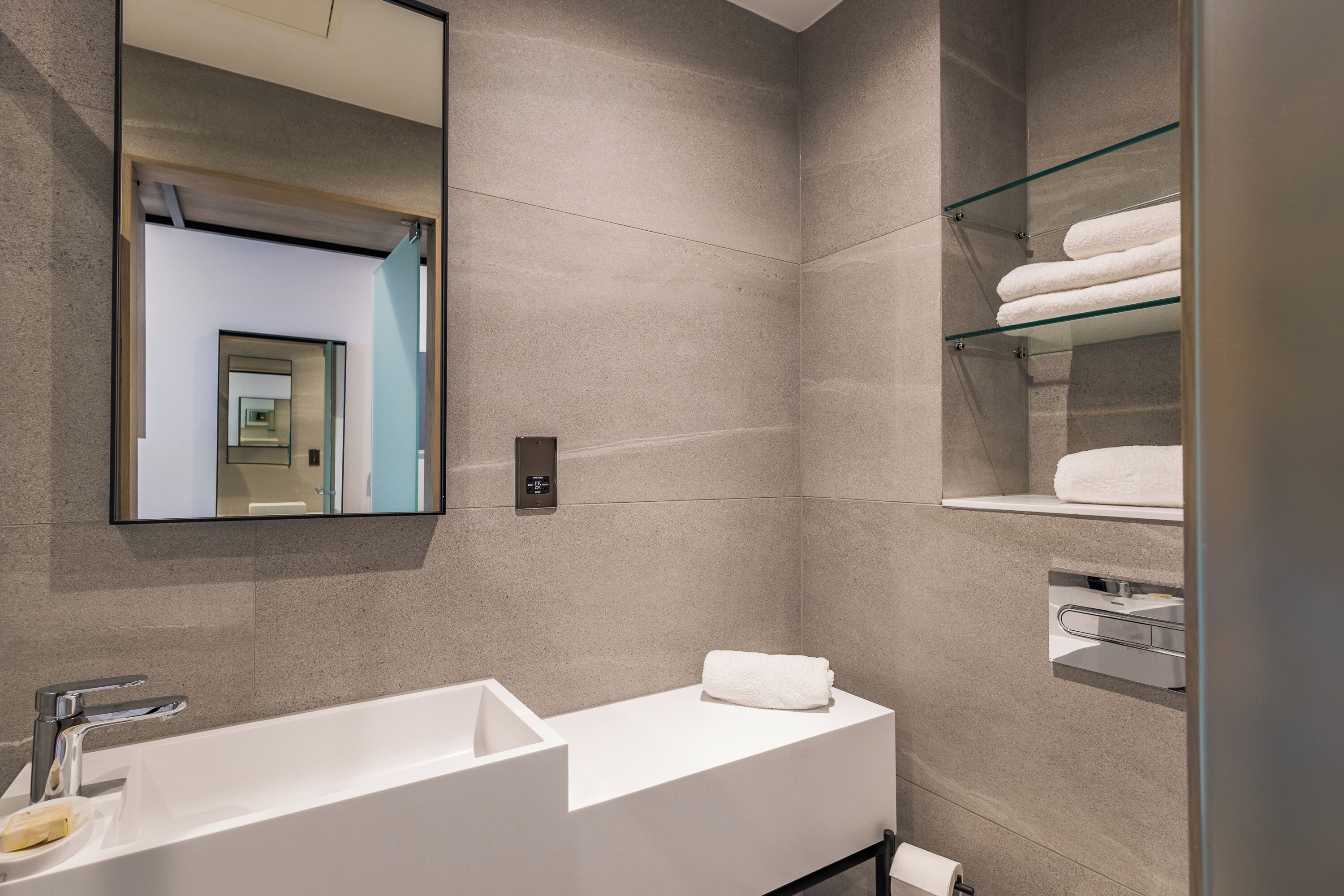 A modern bathroom with a large mirror, a white sink, and a glass shelf with towels.