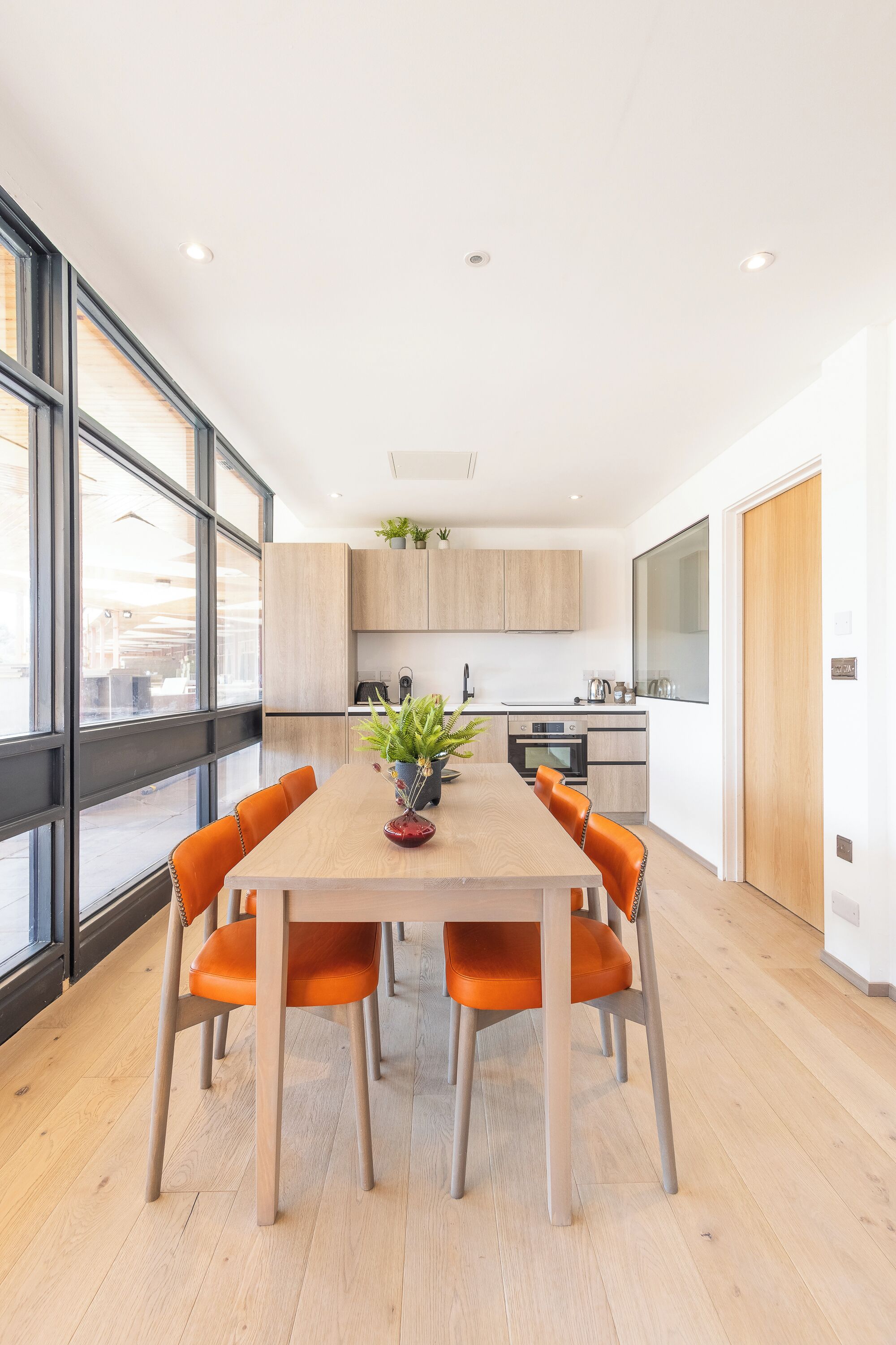 Une cuisine moderne avec des meubles en bois, une table à manger entourée de chaises orange et une grande fenêtre qui laisse entrer la lumière naturelle.
