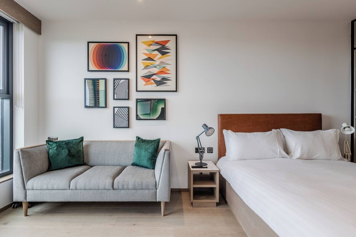 A modern hotel room with a grey sofa, a wooden headboard bed, and artwork on the wall.