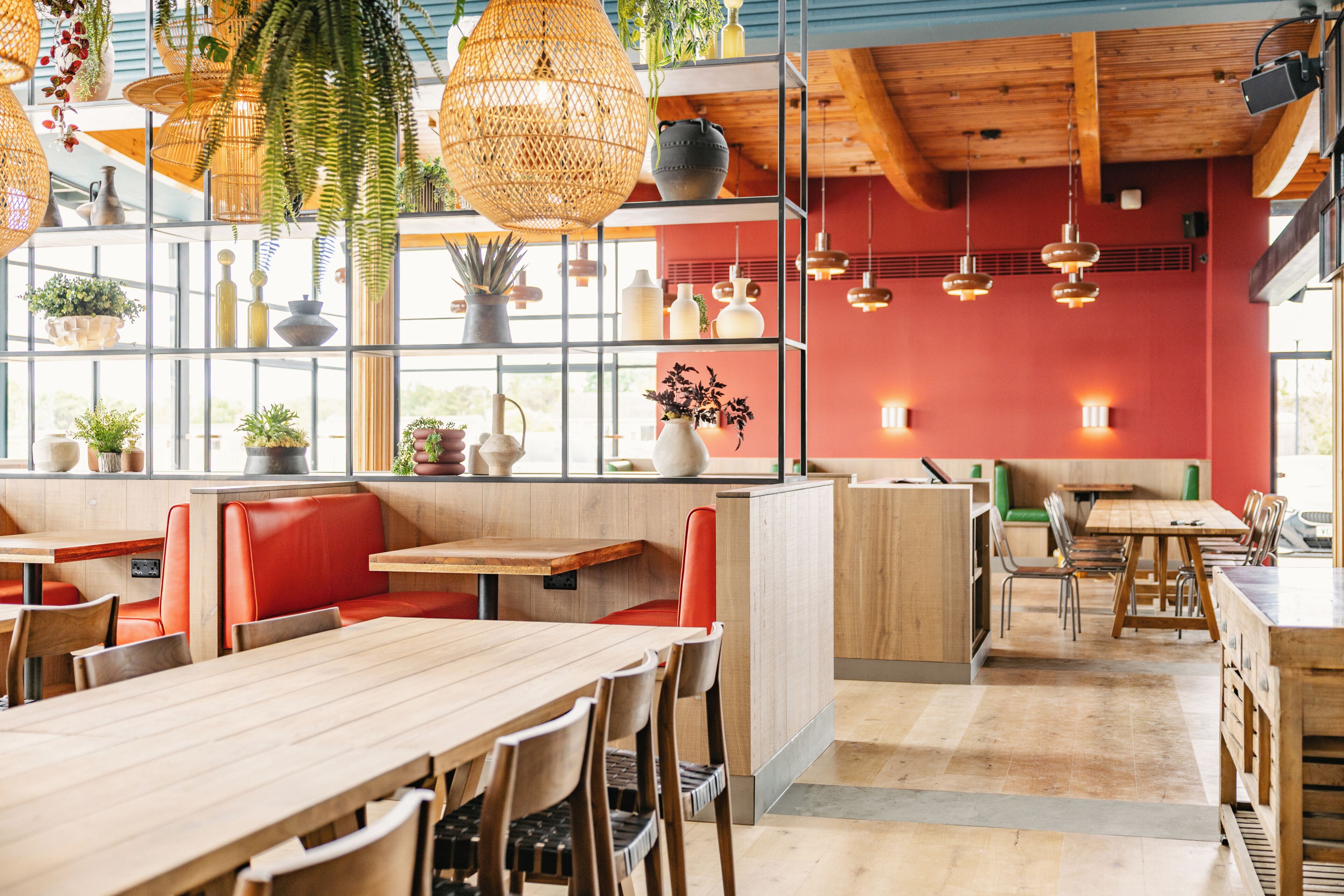 L’intérieur d’un restaurant avec des tables en bois, des sièges rouges, des lampes en osier suspendues et des plantes en pot disposées sur des étagères.