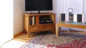 A room with a television on a wooden stand, a bookshelf, a small table with speakers, and a patterned rug. - Ferienhaus Zum Roten Adler in Storkow (Storkow)