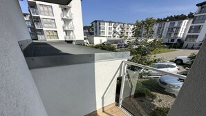 Un balcon doté d’un plancher en bois, d’une rambarde en verre et offrant une vue sur un parking où se trouvent des voitures et, au loin, des immeubles d’habitation.