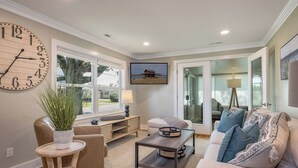 Uma sala de estar com um relógio de parede grande, televisão, sofá, mesa de centro e um vaso de planta.