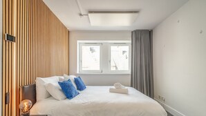 A modern hotel room with a large bed, a bedside table with a lamp, a window with curtains, and a wooden wall panel.