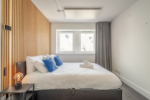 A modern hotel room with a large bed, a bedside table with a lamp, a window with curtains, and a wooden wall panel. - Muttoes Apartments (No 21) (St Andrews)