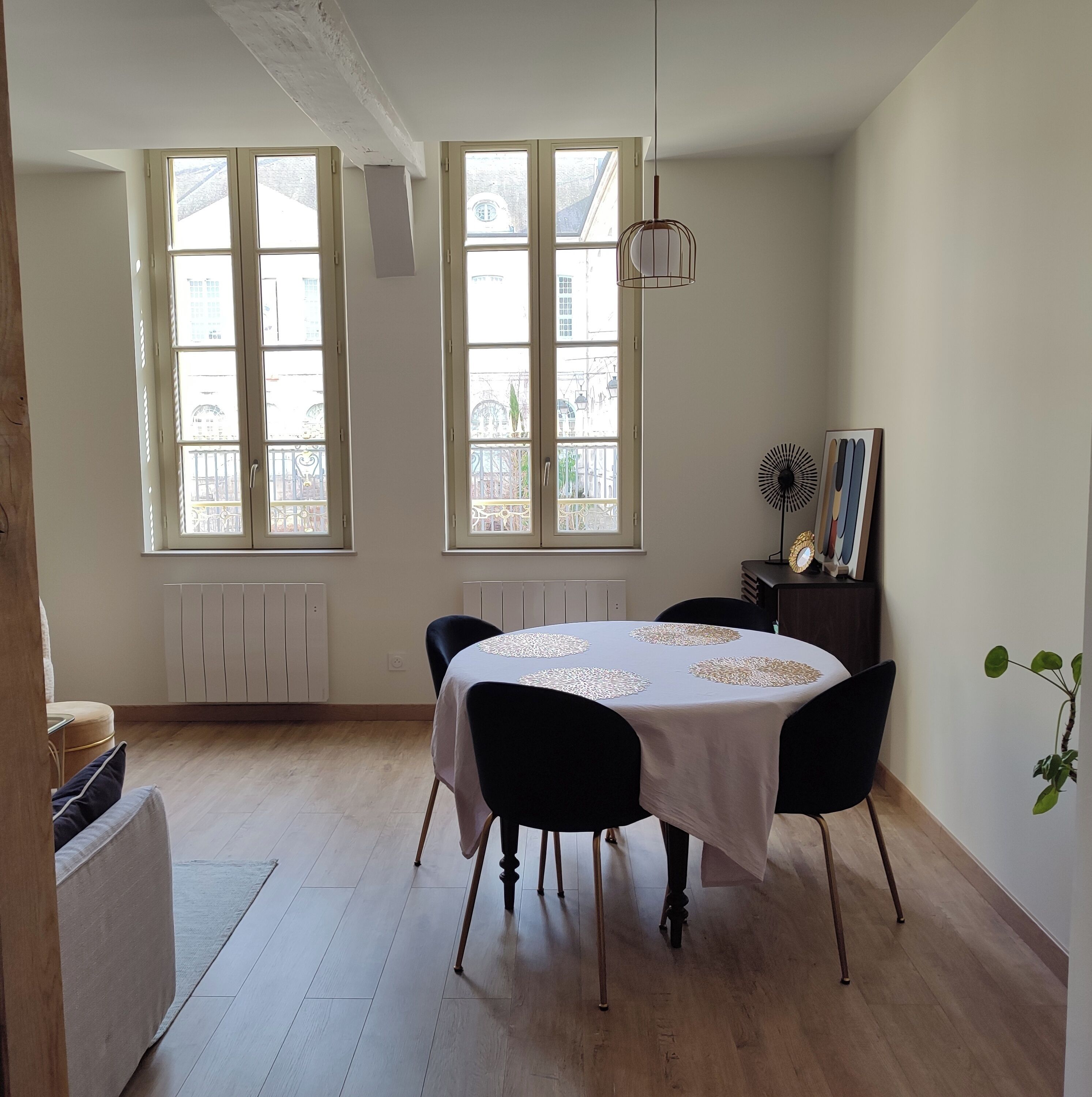 Un comedor con una mesa redonda, sillas negras y un mantel blanco. Hay dos ventanas con marcos blancos y vistas a edificios. Una lámpara colgante cuelga del techo, y una planta pequeña está a la derecha.