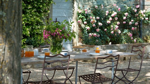 Un área de comedor al aire libre con una mesa de madera rústica y sillas de metal, rodeada de exuberante follaje y flores en flor.