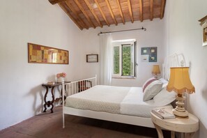Un dormitorio con techo de madera, una cama con ropa de cama blanca, una mesita de noche con una lámpara y una ventana con cortinas.