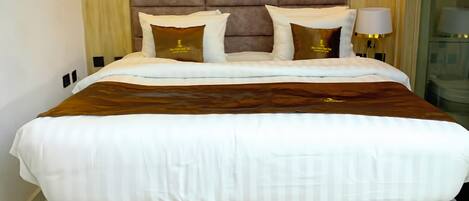 Una cama grande con cabecera acolchada, ropa de cama blanca y dos almohadones decorativos.