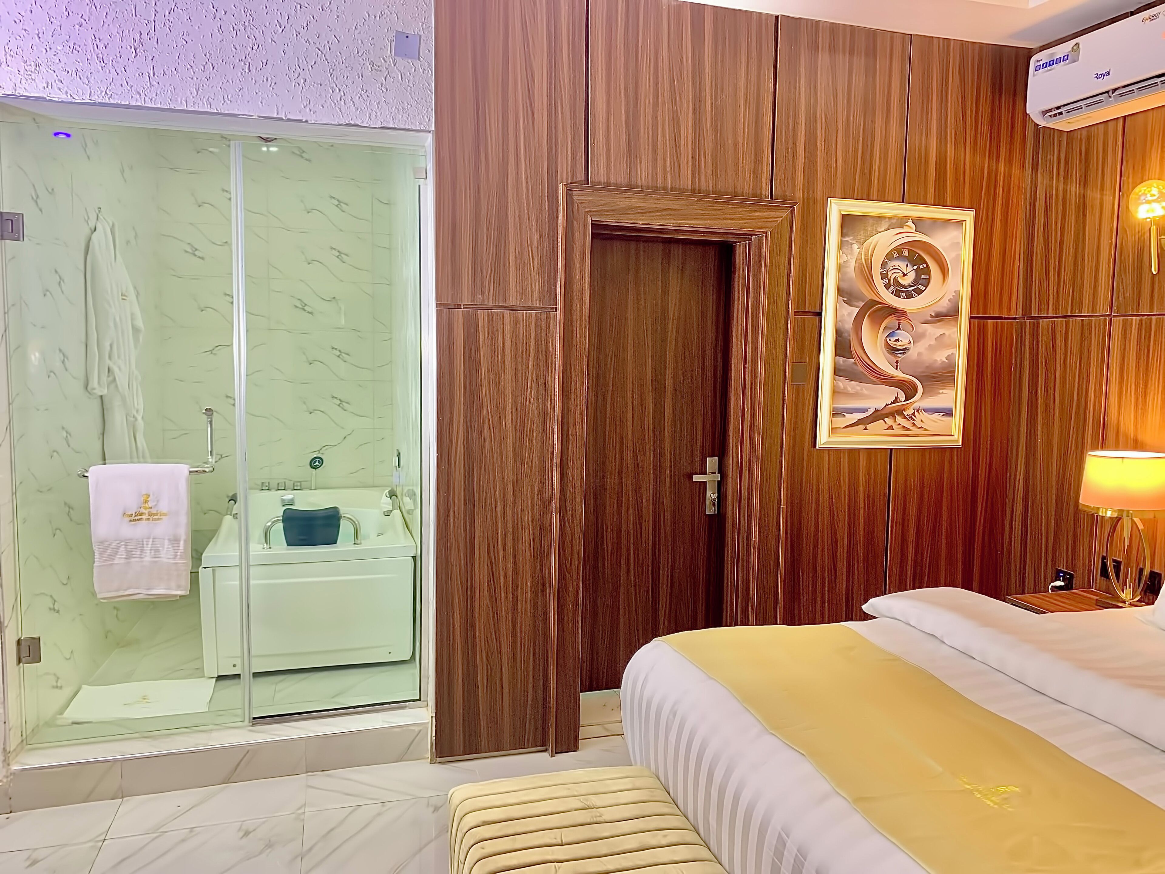 A hotel room with a glass shower, wooden panel walls, a bed with a yellow bedspread, and a wall-mounted clock.