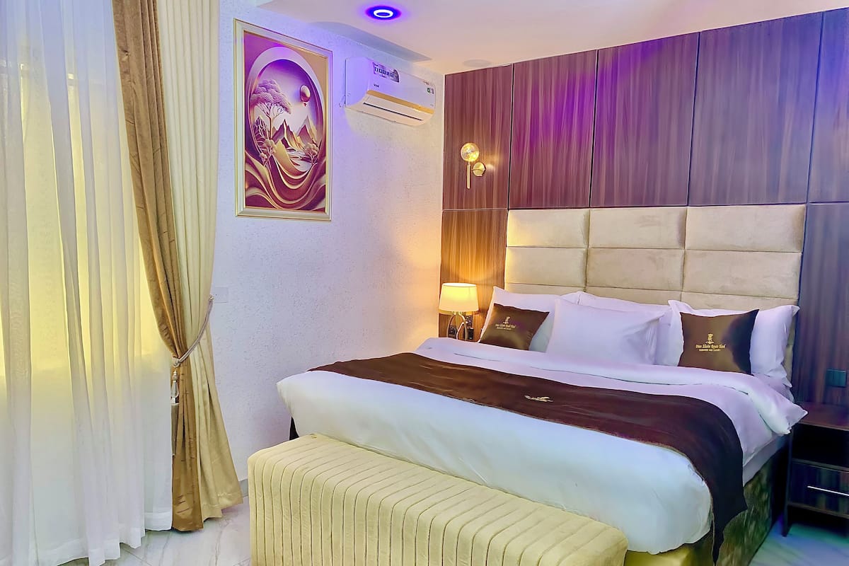 A hotel room with a bed, a headboard, a bedside table with a lamp, and a wall-mounted air conditioner.