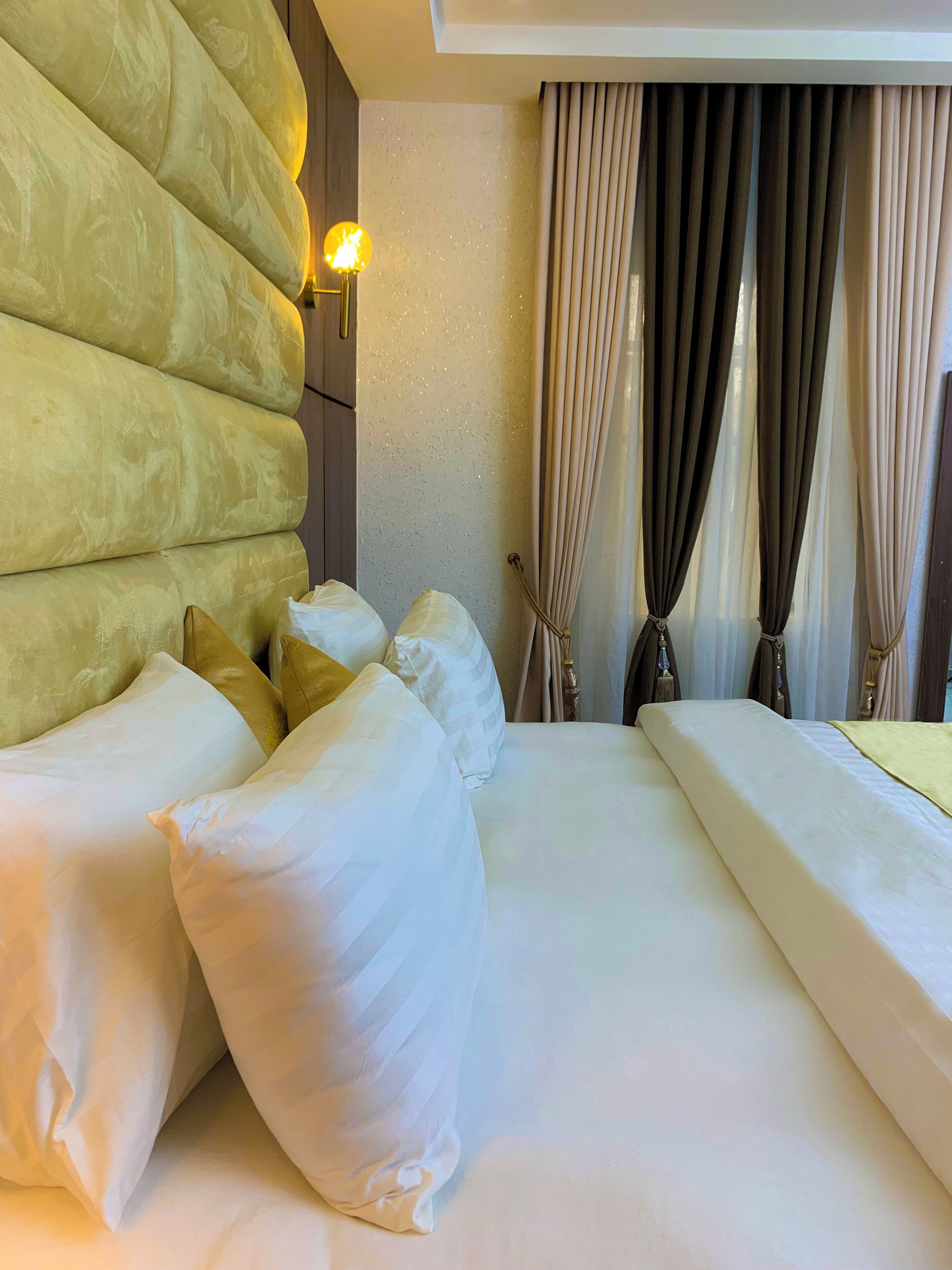 A hotel room with a large bed, multiple pillows, a wall-mounted light, and a window with curtains.
