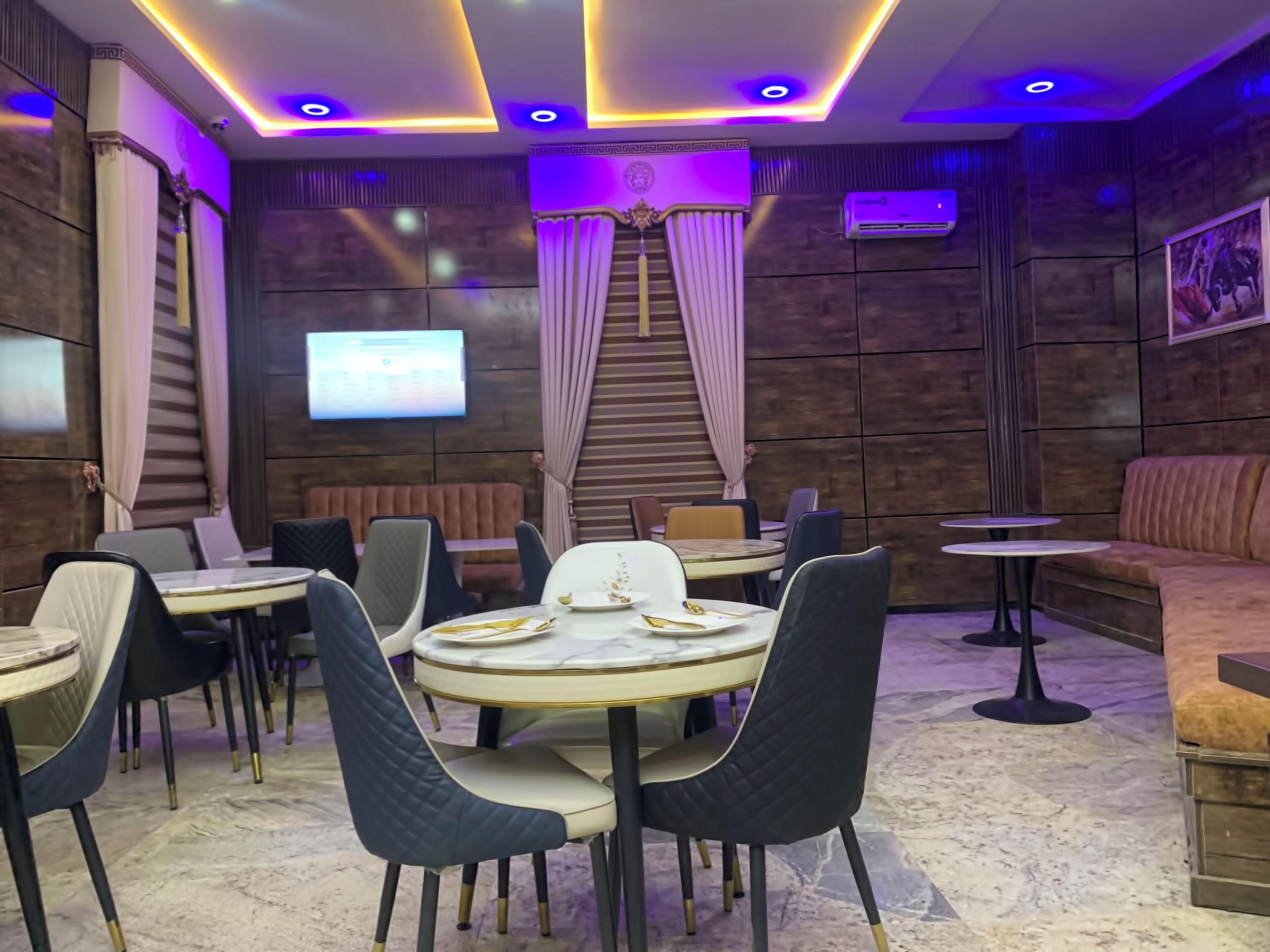 A modern restaurant interior with round tables, cushioned chairs, a sofa, and a TV screen.