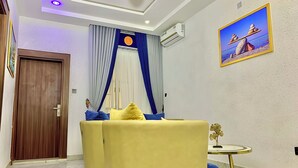 Une chambre d’hôtel avec une porte en bois, un rideau bleu et blanc, un canapé jaune avec des coussins bleus, une petite table ronde et un tableau encadré au mur.