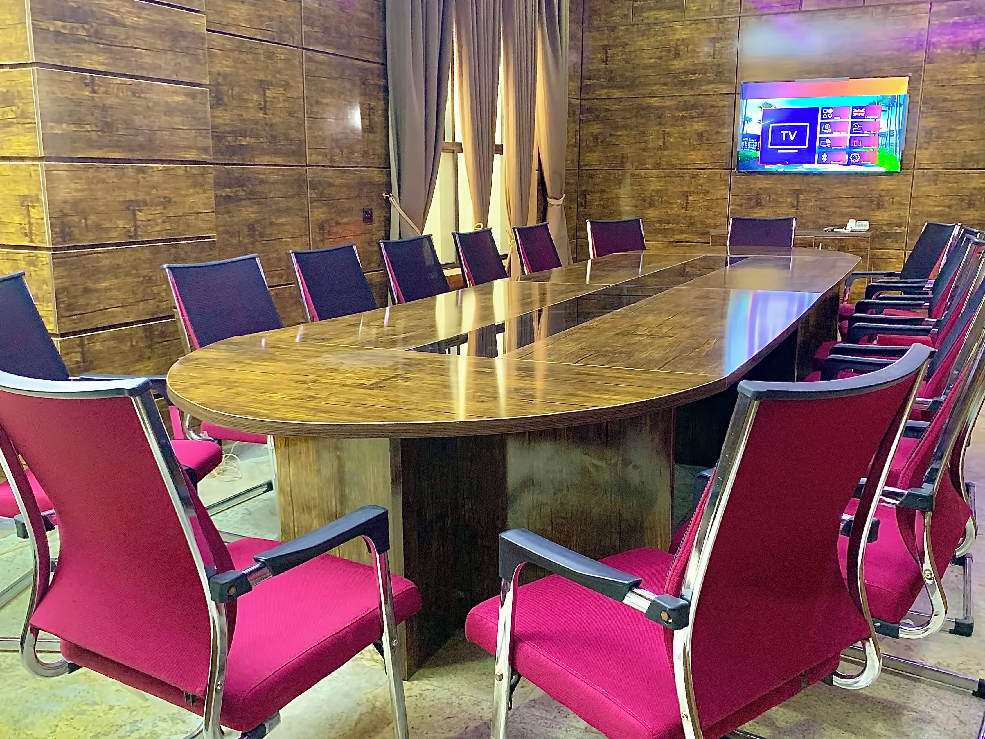 A conference room with a large oval table surrounded by red chairs, a wooden wall, and a TV mounted on the wall.