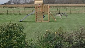 Une structure de jeu en bois, dotée d’un toboggan et d’une échelle d’escalade, se trouve dans un champ herbeux, à proximité d’une table de pique-nique.