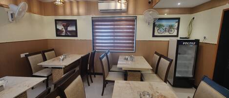 A restaurant interior with wooden paneling, two dining tables with chairs, a refrigerator, and wall-mounted fans.