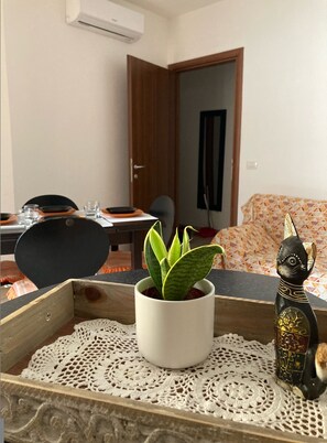 Ein Zimmer mit Esstisch für zwei Personen, einem Blumentopf, einer dekorativen Katzenfigur und einer Holzkiste mit Unterlage.