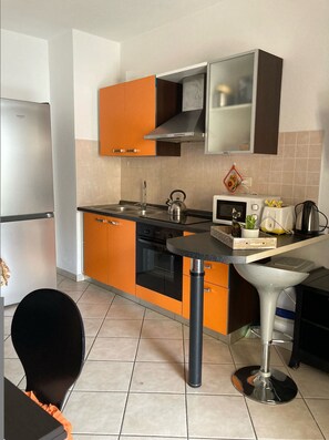 Eine kompakte Küche mit orangefarbenen Schränken, einem Edelstahlkühlschrank, einer Mikrowelle und einem Wasserkocher auf der Arbeitsfläche.
