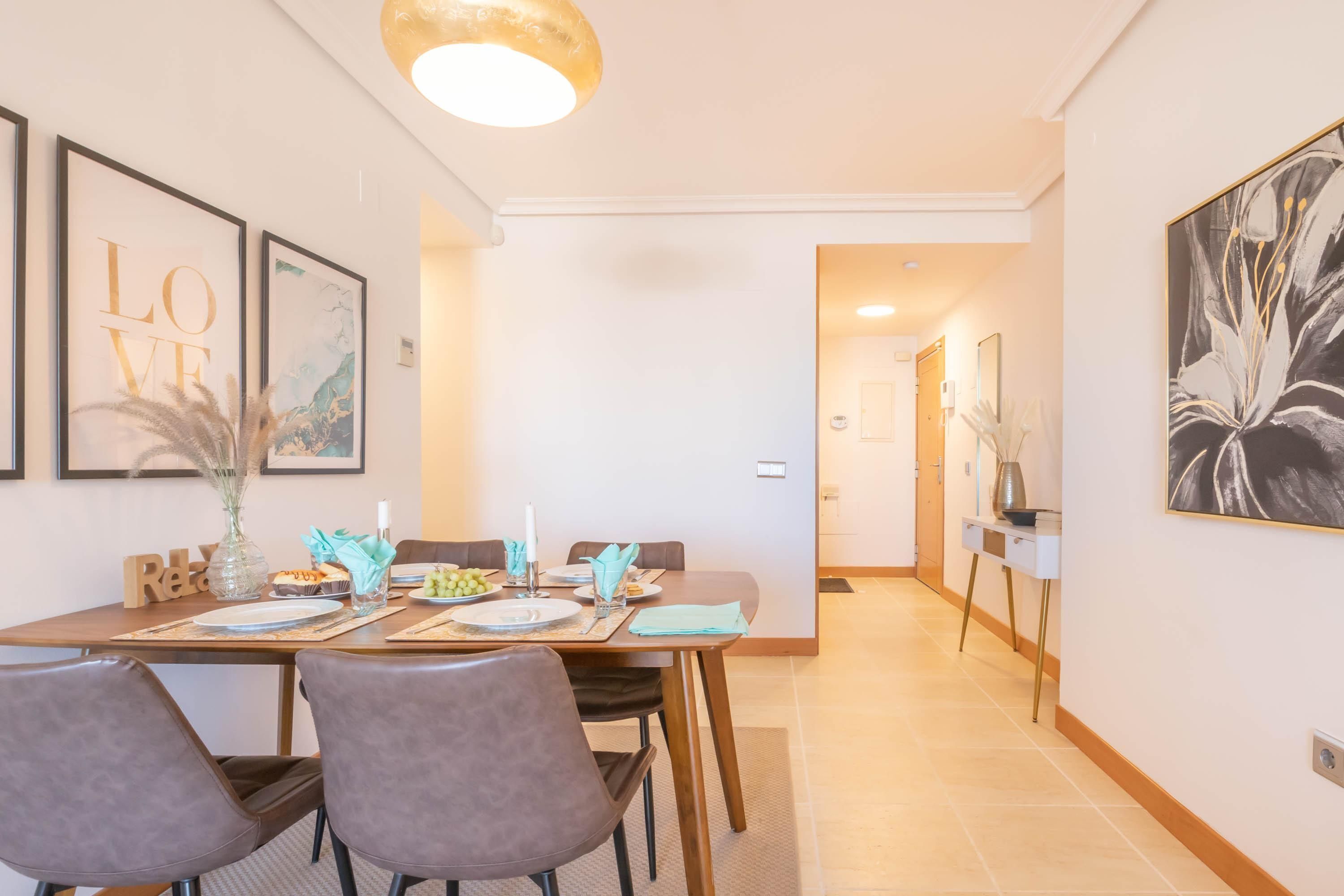 Un coin repas avec une table en bois dressée pour quatre personnes, des œuvres d’art encadrées accrochées aux murs et un couloir menant à une autre pièce.