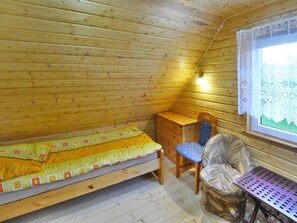 A small, cozy room with a wooden sloped ceiling, a single bed with a colorful blanket, a wooden desk, a chair, and a window with lace curtains. - Large Holiday Home With a Lake View (Malechowo)