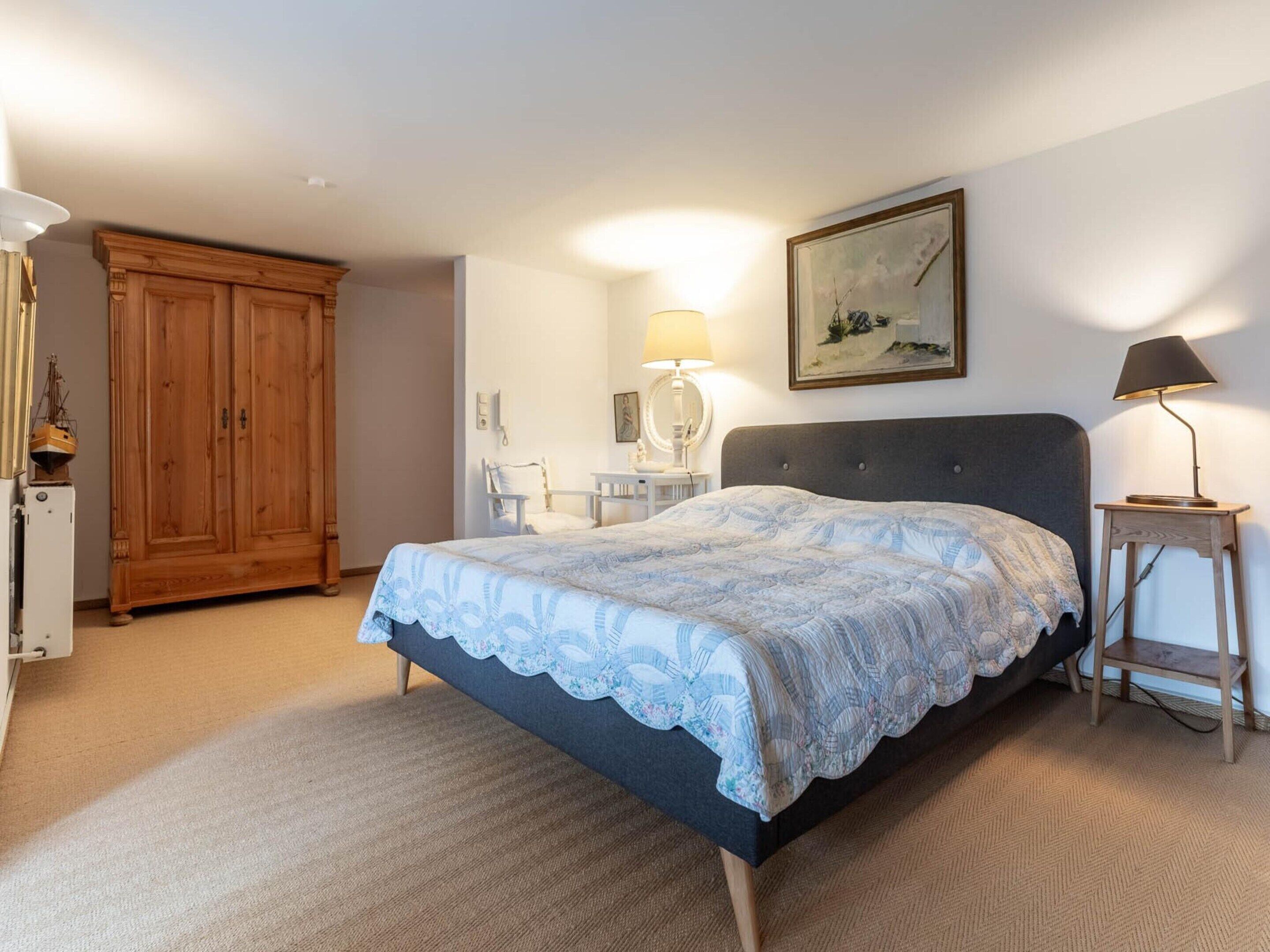 Ein Schlafzimmer mit einem Bett, einem Holzschrank, einem Schreibtisch mit Stuhl, einer Lampe und einem Bild an der Wand.