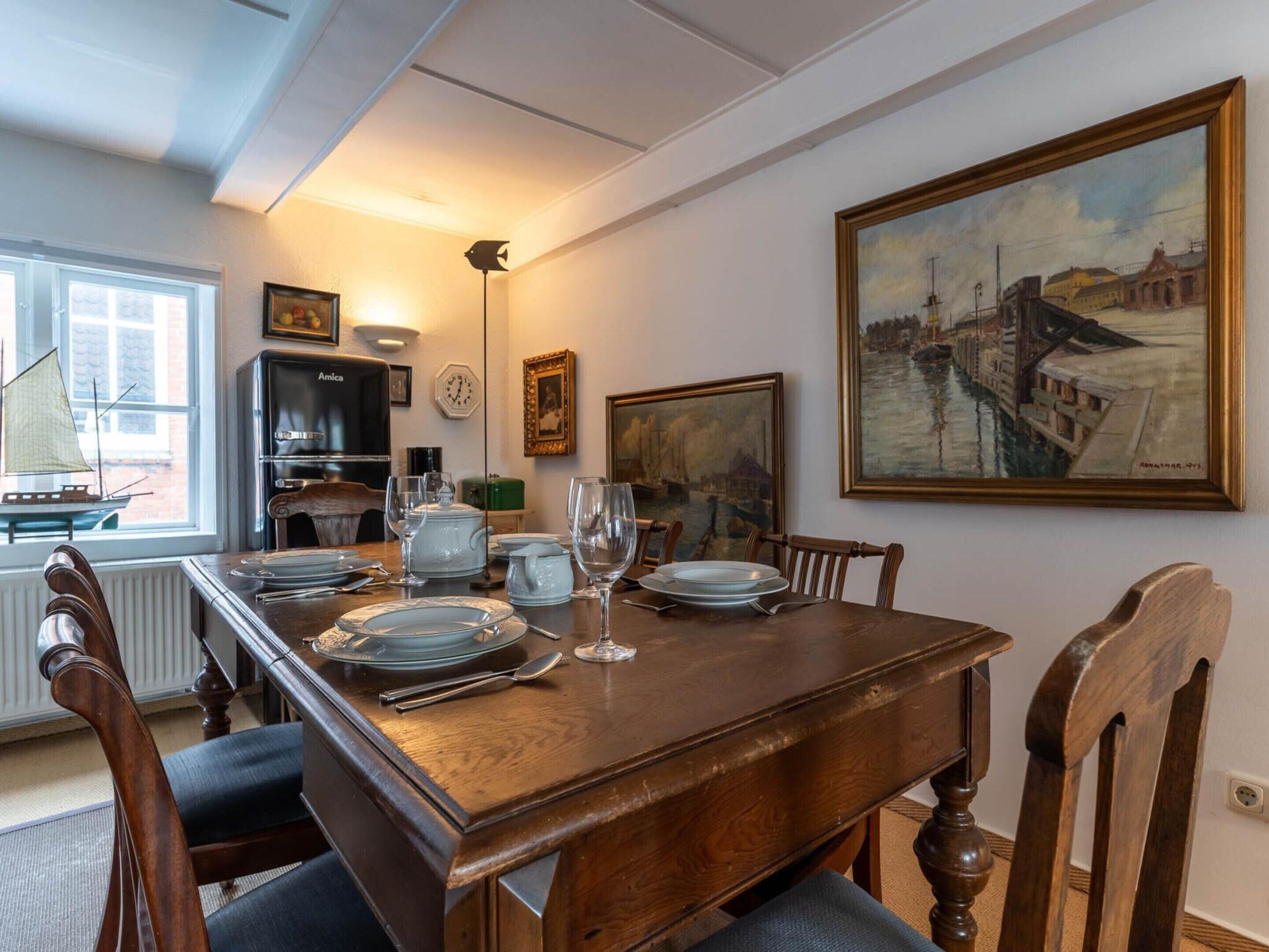 Uma sala de jantar com mesa de madeira posta para quatro, cadeiras, relógio e um quadro na parede.