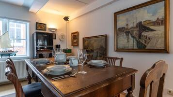 Uma sala de jantar com mesa de madeira posta para quatro, cadeiras, relógio e um quadro na parede.