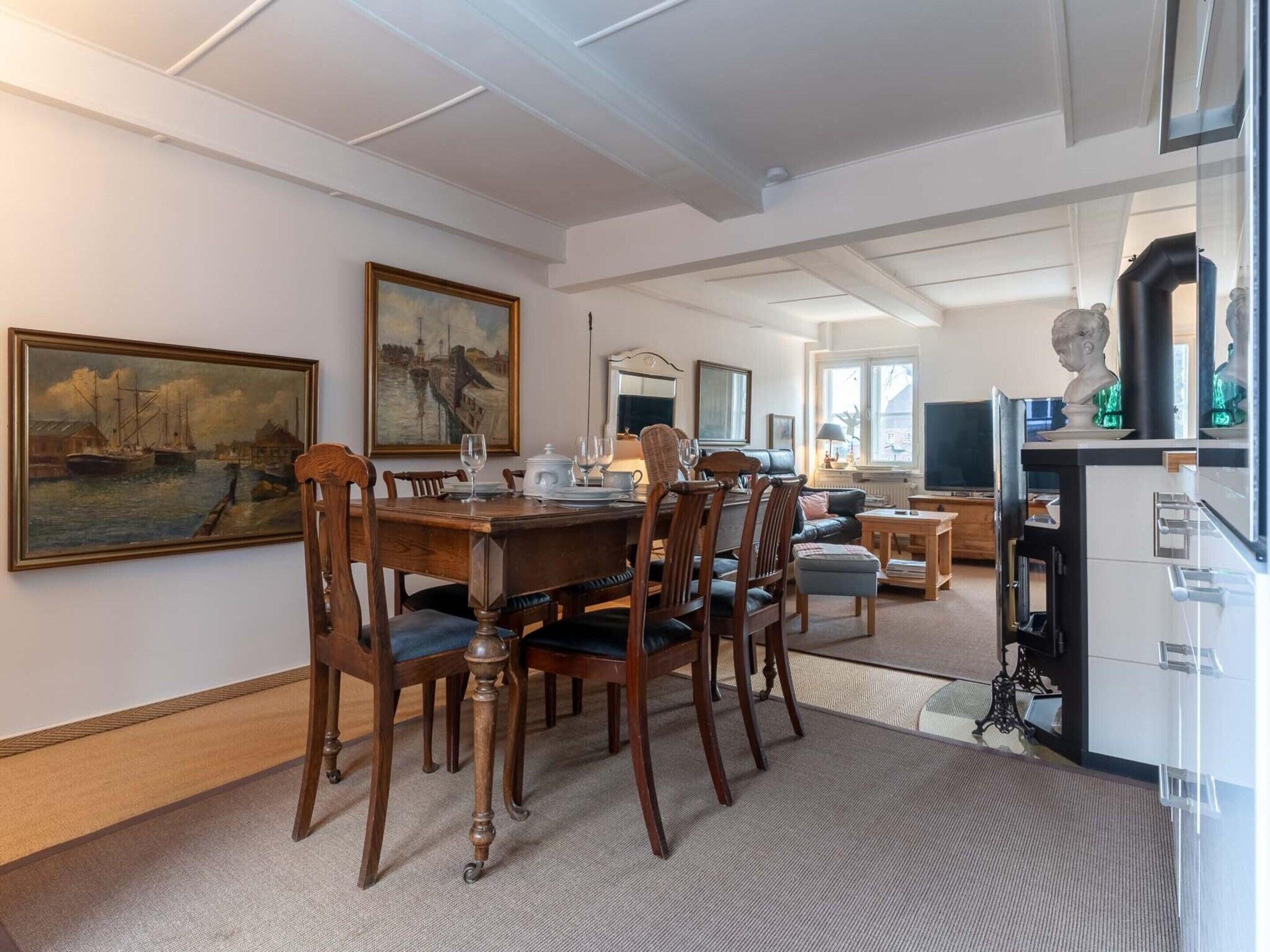 A dining area with a wooden table and chairs, a painting on the wall, and a television in the background.