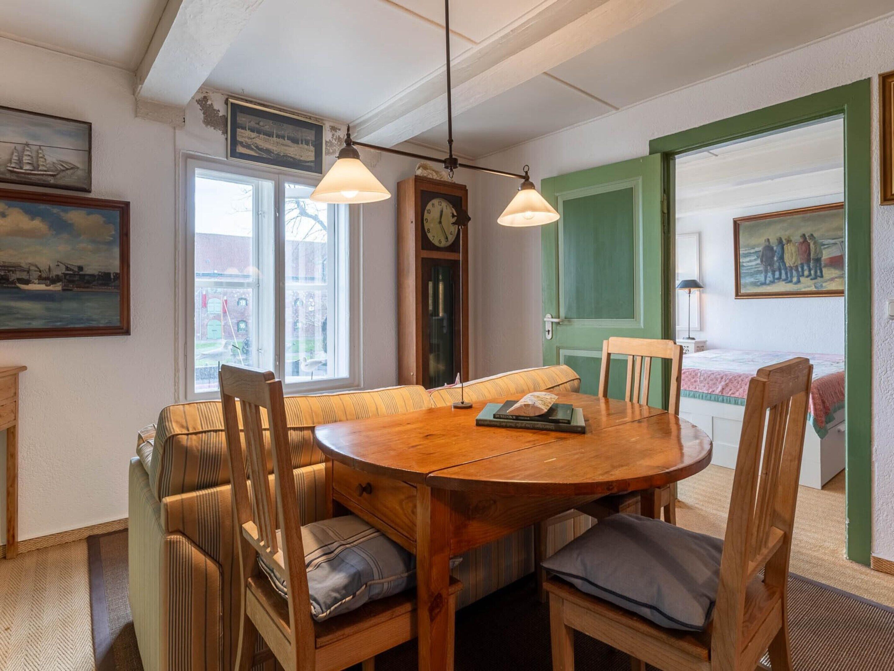 A cozy dining area with a wooden table, chairs, and a bench. There is a clock on the wall, a window with a view, and a door leading to another room.