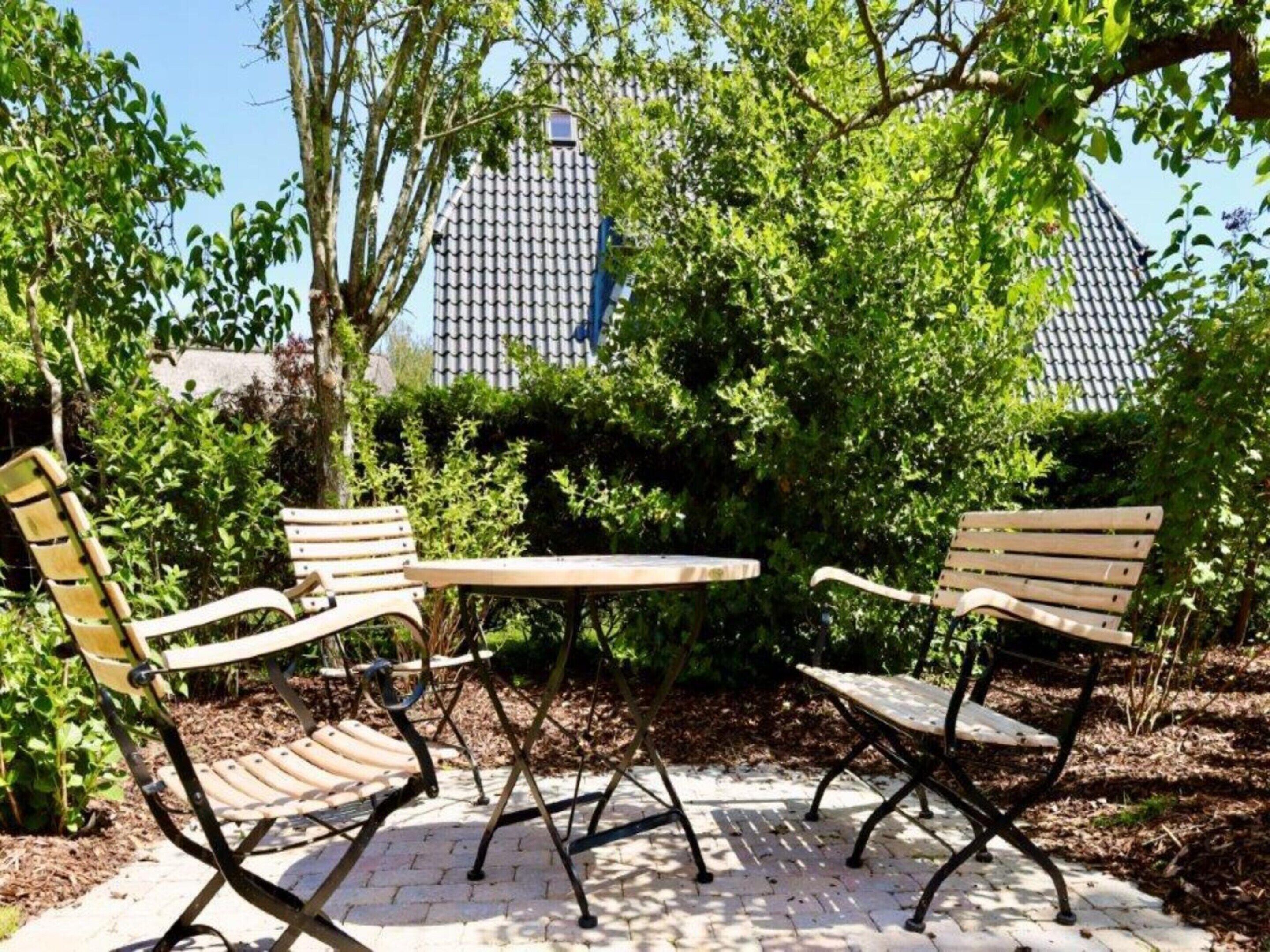 An outdoor seating area with two wooden chairs and a table, surrounded by greenery and a tiled roof in the background.