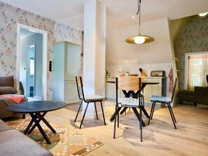 Sala de estar com sofá, mesa de centro e quatro cadeiras dispostas ao redor. Na parte de trás, há uma cozinha com mesa e cadeiras.