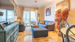 Ein modernes Wohnzimmer mit einer Couch, einem Hocker und einem Essbereich mit Stuhl und Tisch. Es gibt ein großes Fenster mit Vorhängen, eine Hängeleuchte und eine Pflanze.
