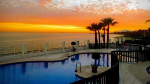 Ein Swimmingpool mit Blick auf den Sonnenuntergang über dem Meer, Palmen und eine umzäunte Liegefläche.