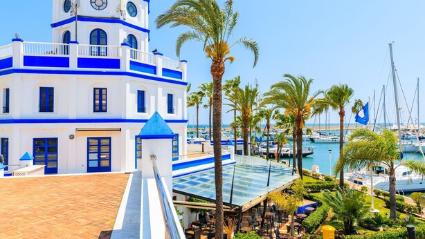 A white building with blue accents, a glass-roofed terrace, and a marina with boats in the background. - Bahia Dorada Complete Villa (Estepona)