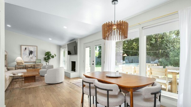 A modern living room with a wooden dining table, light-colored chairs, and a large window offering a view of the outdoors. - Austin Oasis – Zilker, Downtown, Pool, 3 Bedroom (Austin)