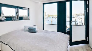 A bedroom with a large bed, a window with a sailboat model, and a view of a marina through an open door. - Houseboat in Egernsund at the Marina Minde (Broager)
