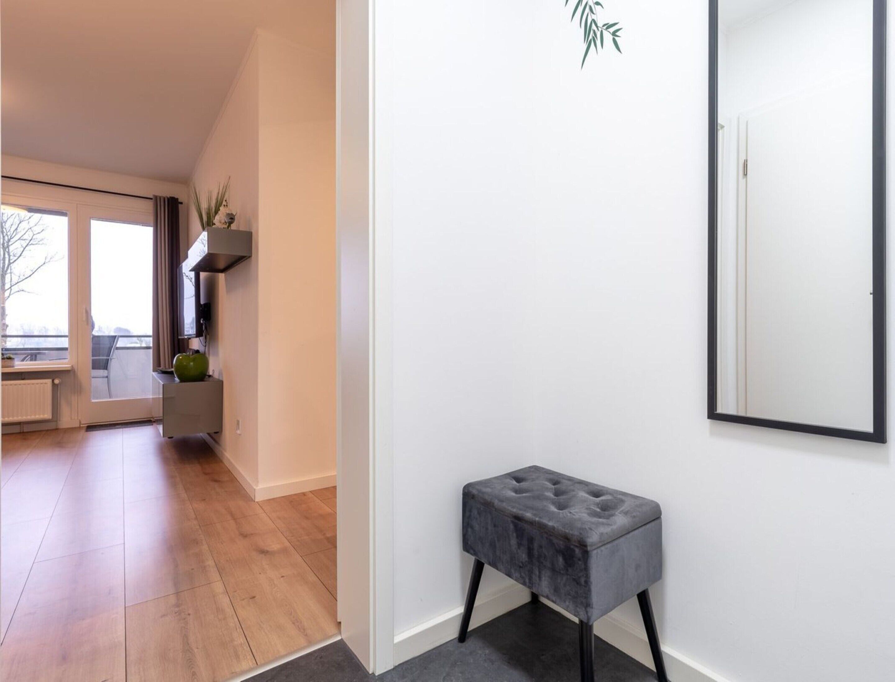 Un interior moderno con un otoman tapizado gris, un espejo grande y vistas a un balcón a través de una puerta de cristal.
