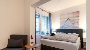 A bedroom with a bed, a chair, a small table, and a window with a view of a brick building. - Ferienwohnung mit Balkon in Westerdeichstrich (Westerdeichstrich)