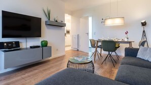 A modern living room with a flat-screen TV, grey sofa, dining table with chairs, and a small round table with a magazine. - Ferienwohnung mit Balkon in Westerdeichstrich (Westerdeichstrich)