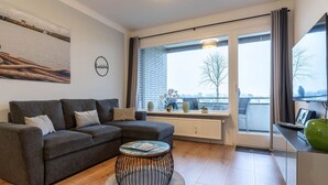 A modern living room with a grey sectional sofa, a round coffee table, and a large window with a view of a brick building and trees. - Ferienwohnung mit Balkon in Westerdeichstrich (Westerdeichstrich)