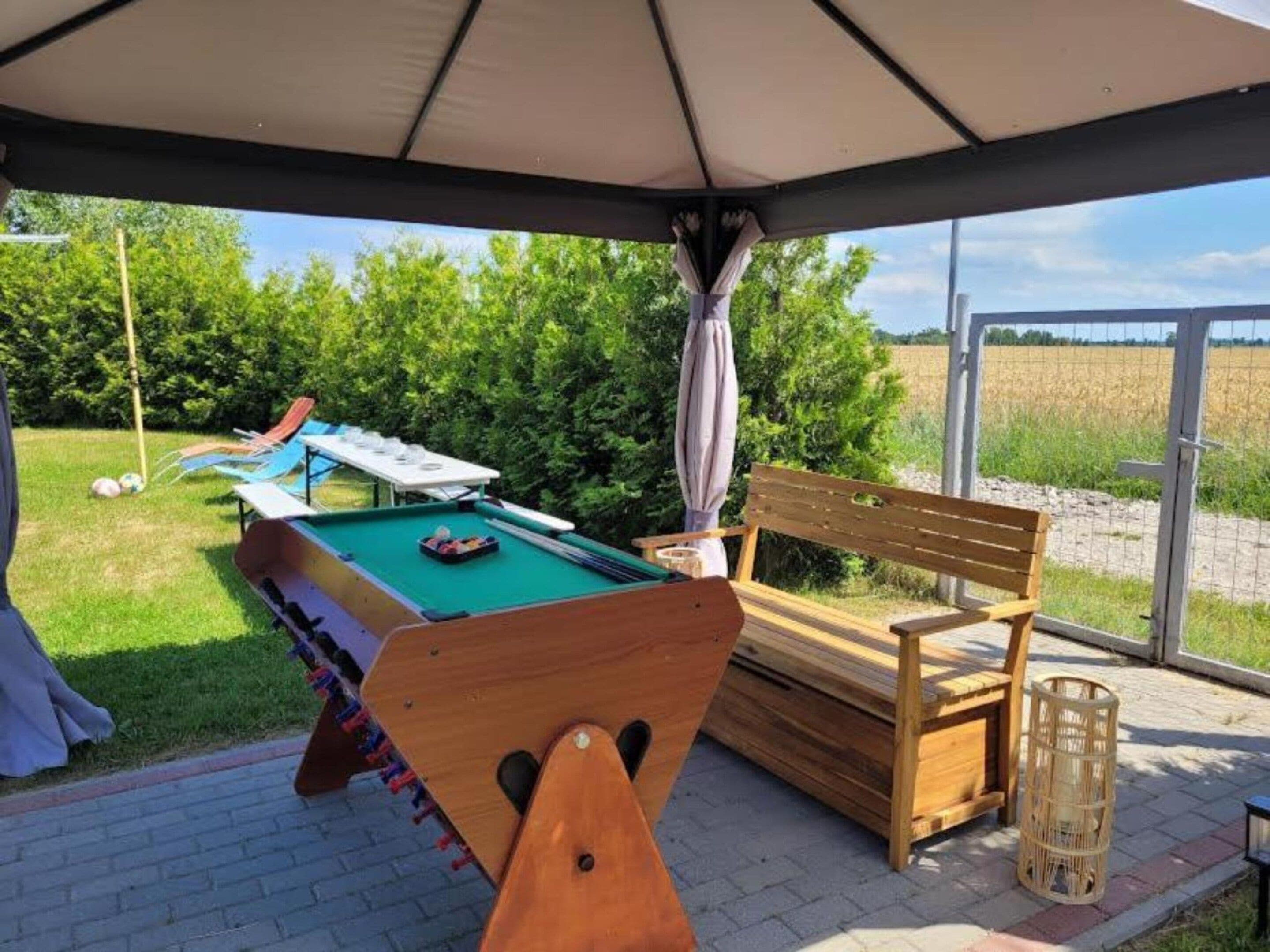 Et udendørs poolområde med en træbænk, et bord med stole og udsigt til et mark.
