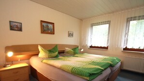 Une chambre à coucher comprenant un lit, deux tables de chevet, une commode, une lampe et deux fenêtres avec des rideaux.