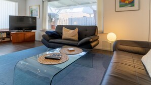 A modern living room with a leather sofa, a glass coffee table, a flat-screen TV, and a large window providing a view of the outdoors. - Comfortable Holiday Apartment in St. Peter Ording (St. Peter-Ording)