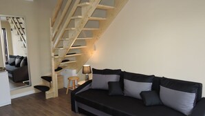 A modern living room with a wooden staircase, a black sofa with grey cushions, a small wooden stool, and a lamp. - Holiday House Near Emerald Lake Wapnica (Międzyzdroje)