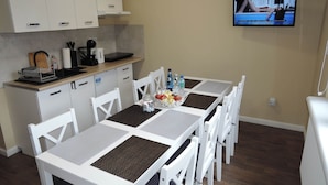 A dining area with a table set for four, surrounded by white chairs. A television is mounted on the wall, displaying a program. - Holiday House Near Emerald Lake Wapnica (Międzyzdroje)