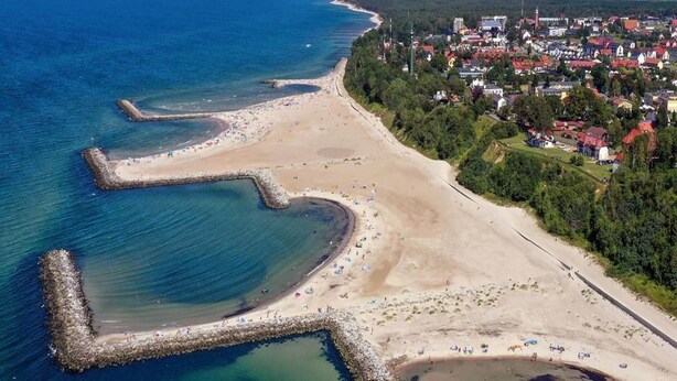 Vista aérea de uma vila costeira com praia arenosa, um cais e edifícios residenciais.