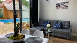 Una sala de estar moderna con un sofá gris, una mesa de comedor con un frutero y un control remoto, y vistas a una piscina exterior.