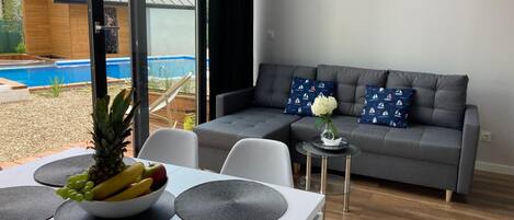 Un salon moderne avec un canapé gris, une table à manger sur laquelle se trouve un saladier rempli de fruits et une télécommande, et une vue sur une piscine à l’extérieur.